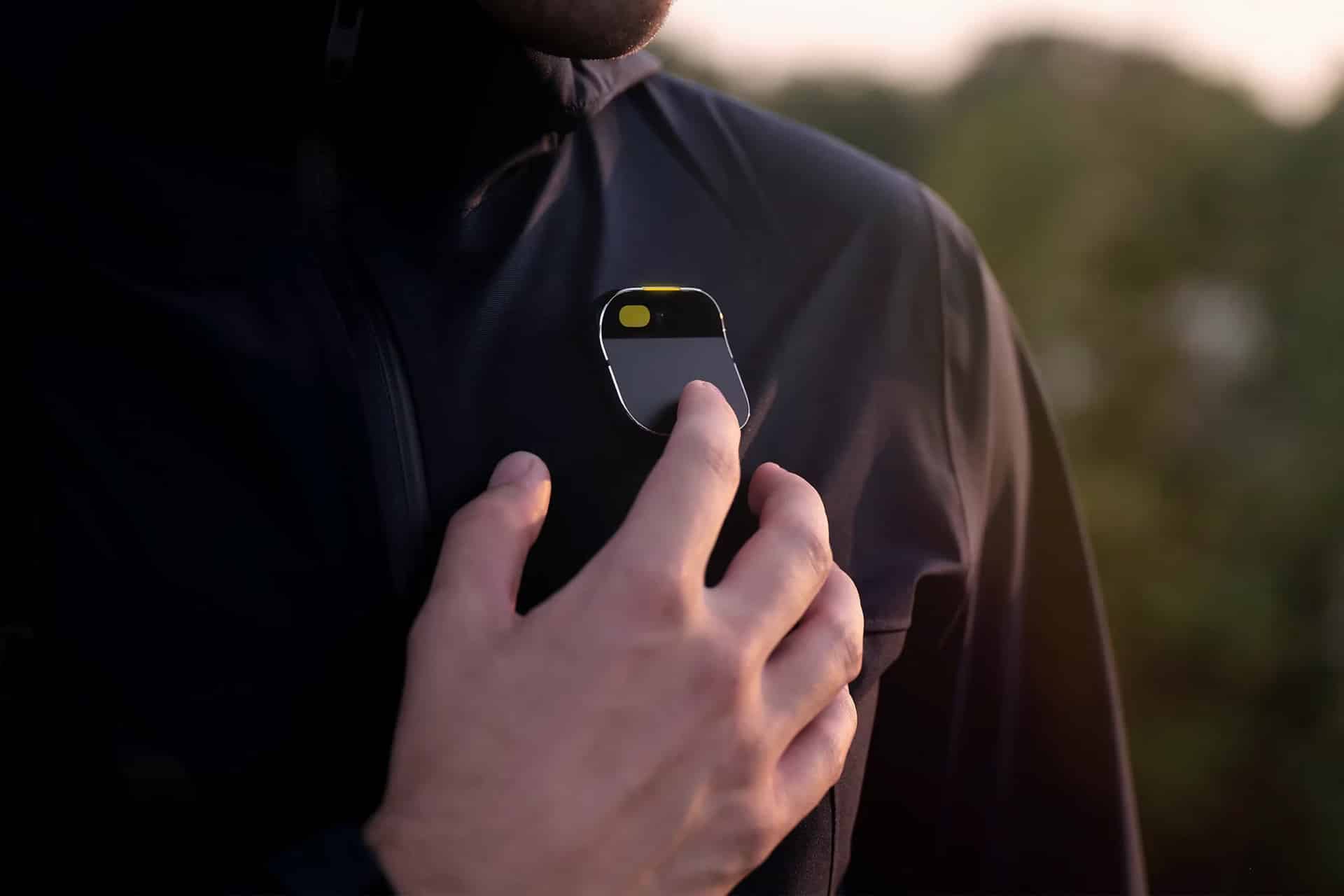 A person wearing a dark jacket attaches a small, sleek, oval-shaped AI Pin with a yellow button to their chest. The background is blurred greenery.