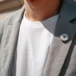 A person wearing a grey blazer and white t-shirt stands outside, with an AI Pin—a small round wearable device—pinned to the blazer's lapel.