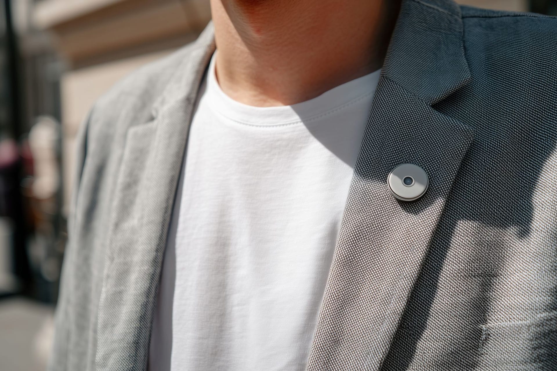 A person wearing a grey blazer and white t-shirt stands outside, with an AI Pin—a small round wearable device—pinned to the blazer's lapel.