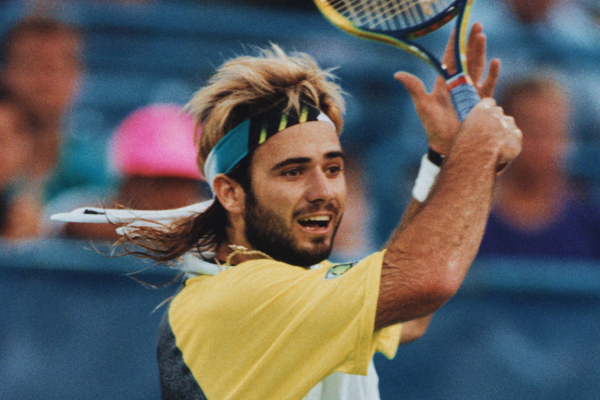 A male tennis player with long hair and a colorful headband, reminiscent of Andre Agassi, prepares to hit a backhand shot during a match in his yellow and white shirt. The blurred background reveals an audience in the stadium.