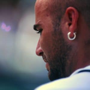A close-up side view of a bald man with a hoop earring, a trimmed beard, and a white shirt, looking down and slightly away from the camera against a blurred background, reminiscent of Andre Agassi's iconic style.