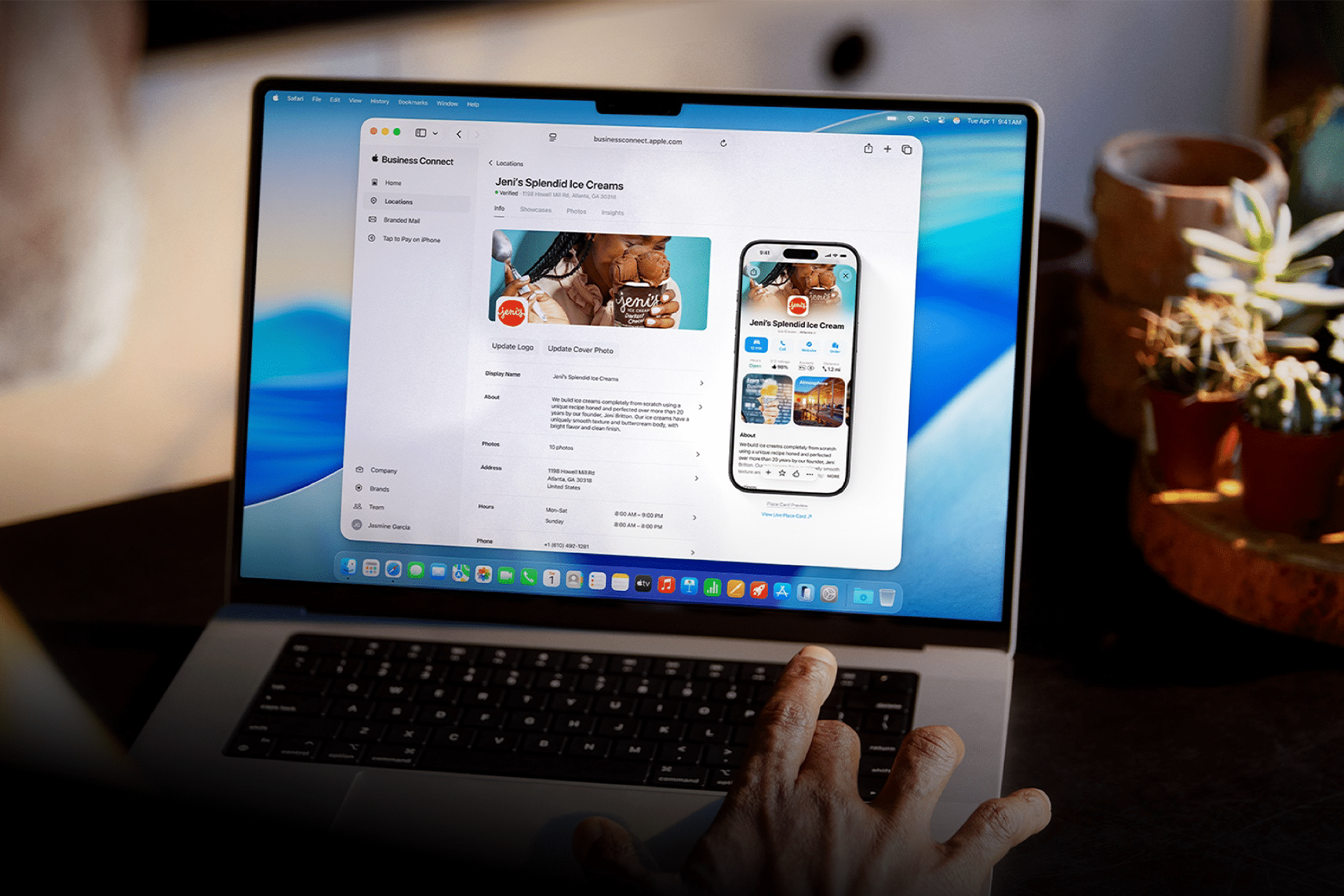 A person’s hand rests on a laptop displaying a business management dashboard with an ice cream brand’s app page and Apple Business Connect, featuring images of ice cream and an app preview on a virtual smartphone. Potted plants are in the background.
