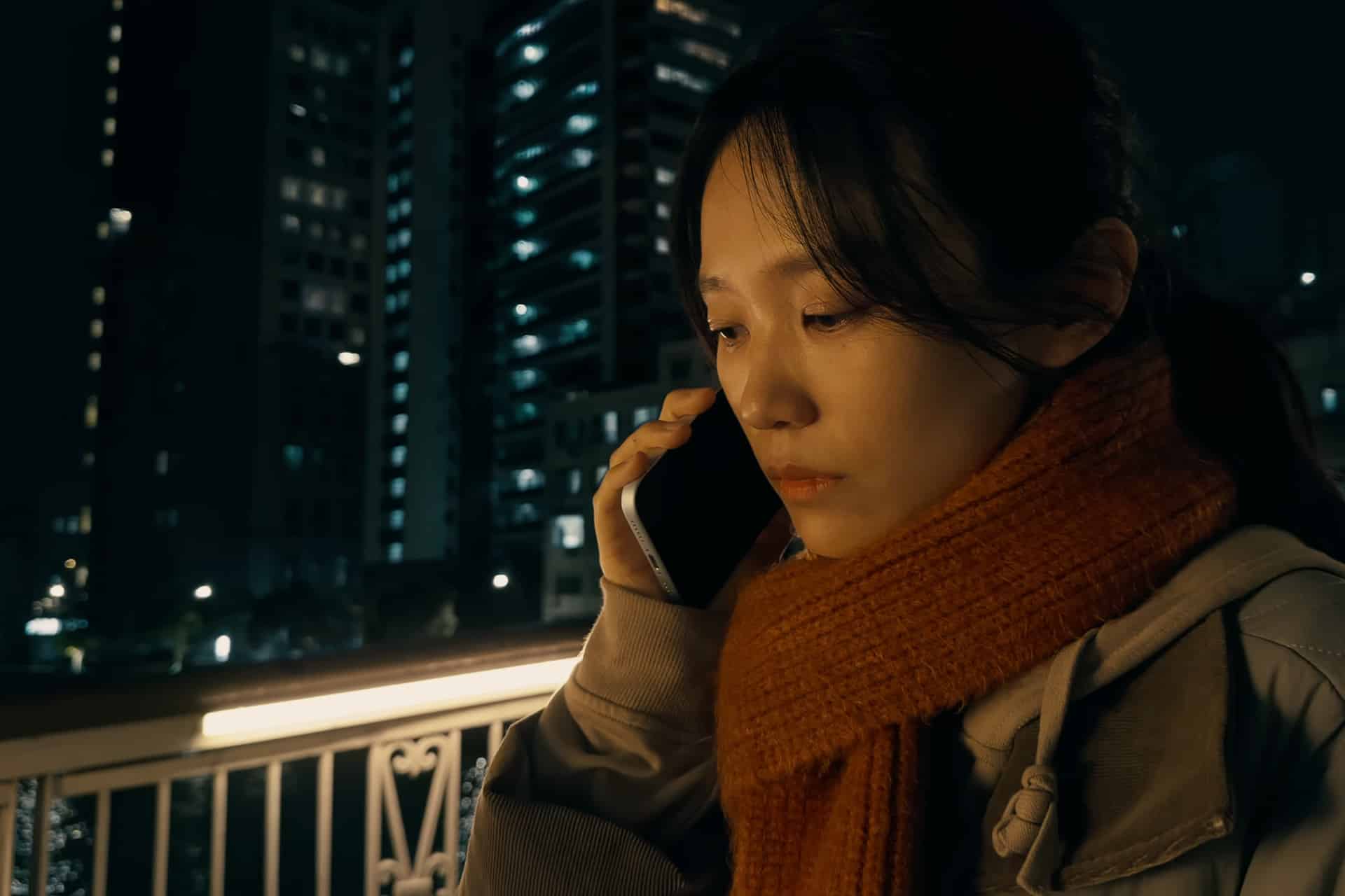 A woman wearing an orange scarf and gray jacket stands on a city bridge at night, talking on her phone. Tall buildings with lit windows form a blurred background. Shot on iPhone.