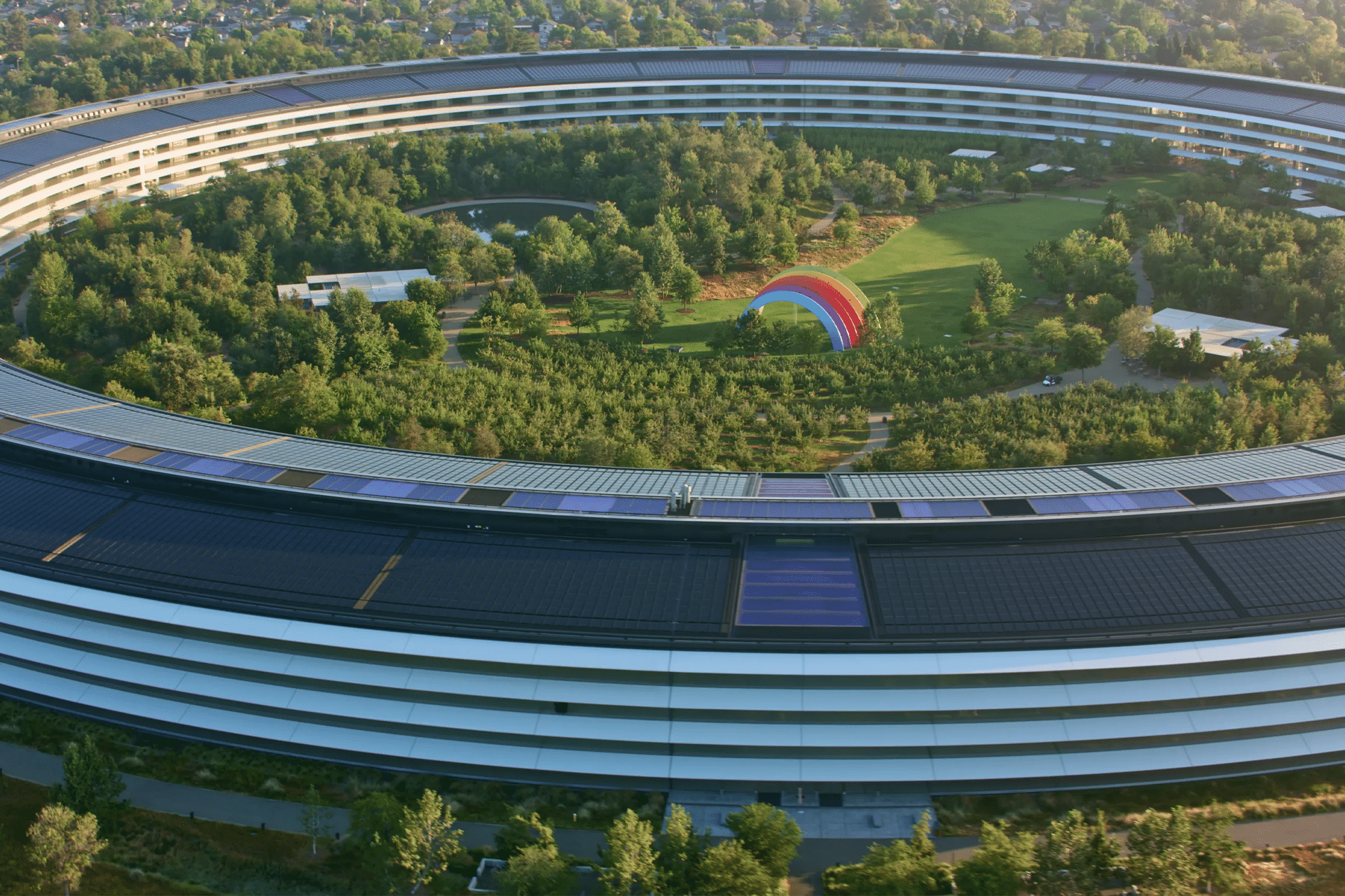 Aerial view of Apple Park, a circular building surrounded by trees, with a large colorful rainbow structure and green lawns inside the central courtyard—a testament to innovation fueled by notable Apple acquisitions.