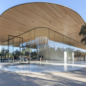 A modern building with a large, curved wooden roof, floor-to-ceiling glass walls, and open spaces—reminiscent of the sleek design seen in Apple Acquisitions. People relax inside and out, with trees surrounding the structure on a sunny day.