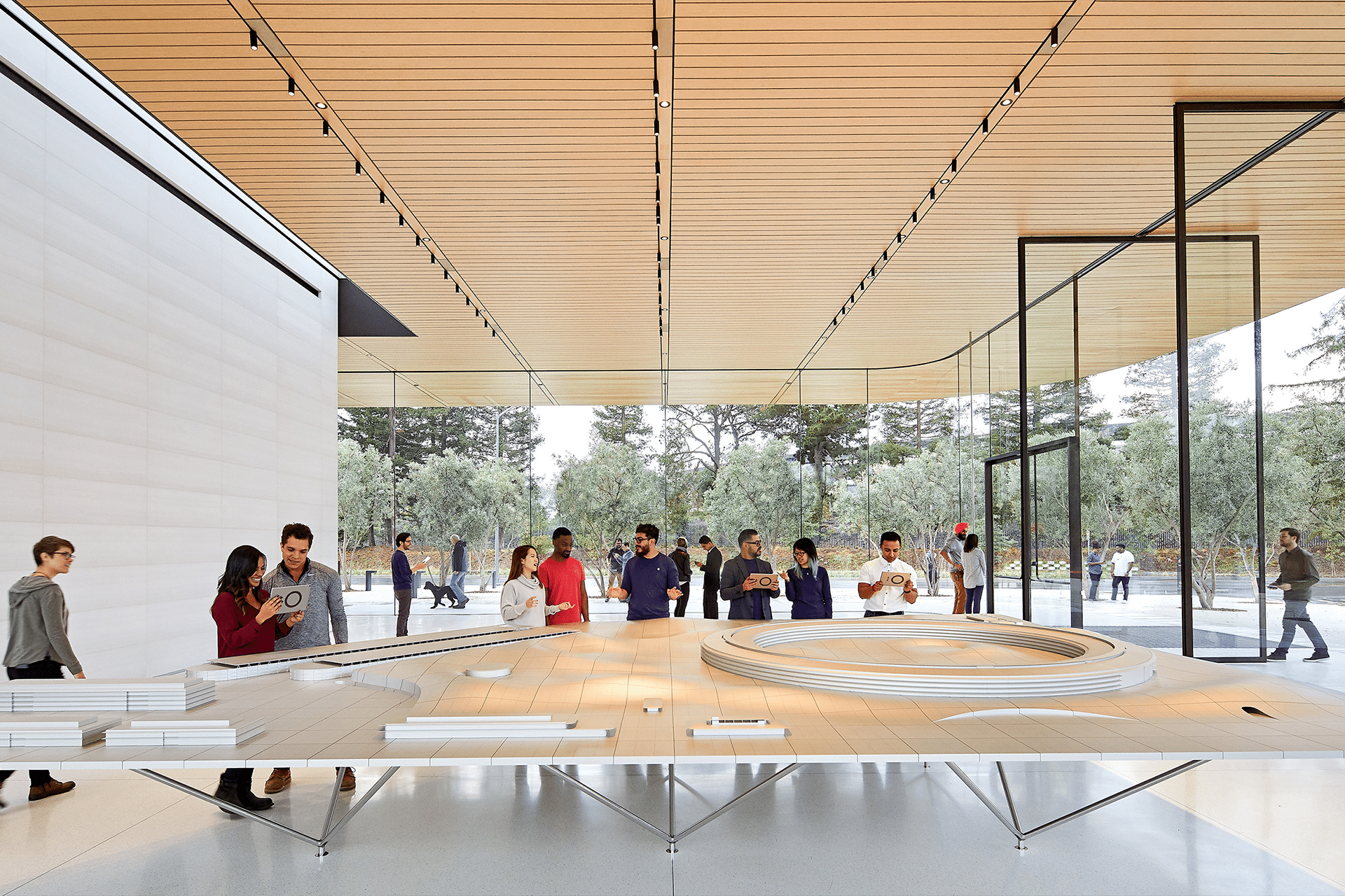 A group of people stands around a large architectural model inside a modern building with tall glass walls and wood ceiling, discussing potential apple Acquisitions as some observe the model closely, while others talk or take photos.