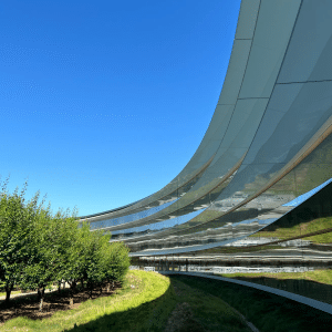 A modern curved glass building, reminiscent of Apple acquisitions, reflects green trees and grass under a clear blue sky, creating a sleek and futuristic architectural scene.