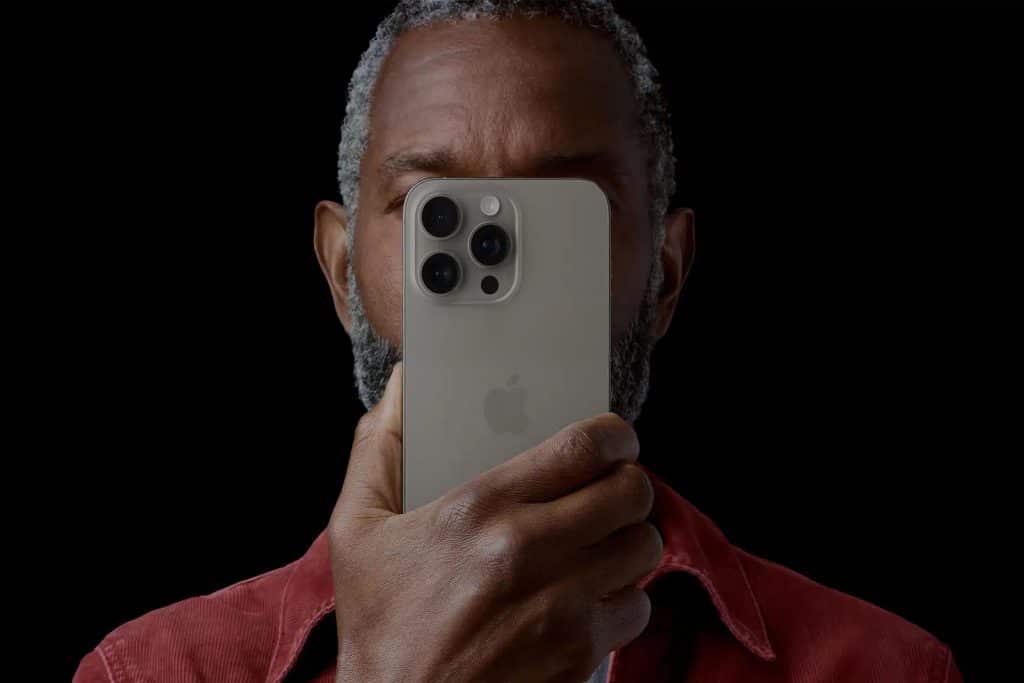 A person with short gray hair and a beard holds a silver iPhone close to their face, partially covering it. The black background makes the phone and person stand out—reminding us how important it is to protect iPhone photos and privacy.