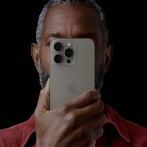 A person with short gray hair and a beard holds a silver iPhone close to their face, partially covering it. The black background makes the phone and person stand out—reminding us how important it is to protect iPhone photos and privacy.