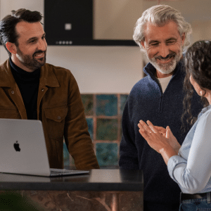 Three people stand around a kitchen counter with a laptop, smiling and engaged in conversation, as Apple Small Business Experts foster a casual and friendly atmosphere.