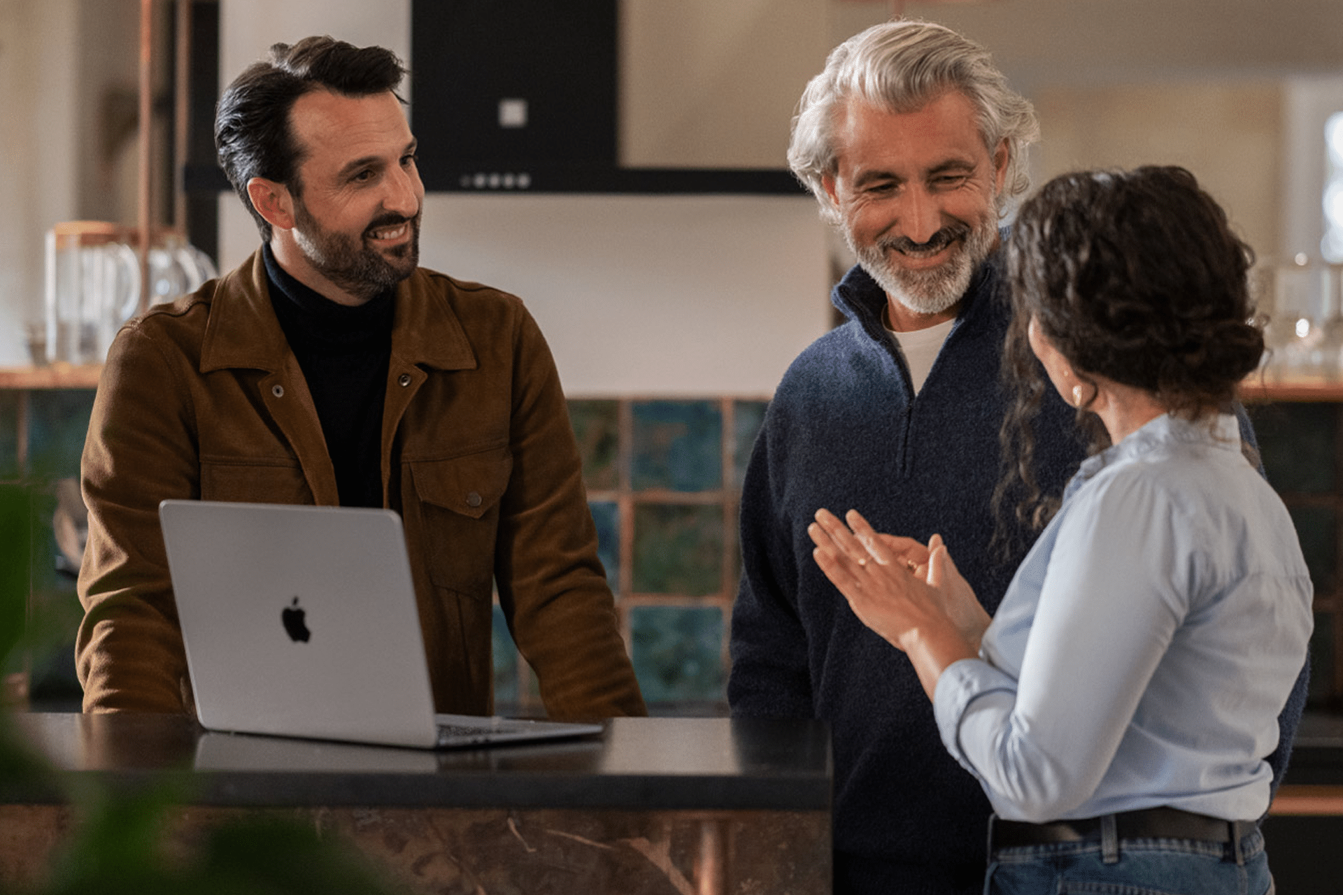 Three people stand around a kitchen counter with a laptop, smiling and engaged in conversation, as Apple Small Business Experts foster a casual and friendly atmosphere.