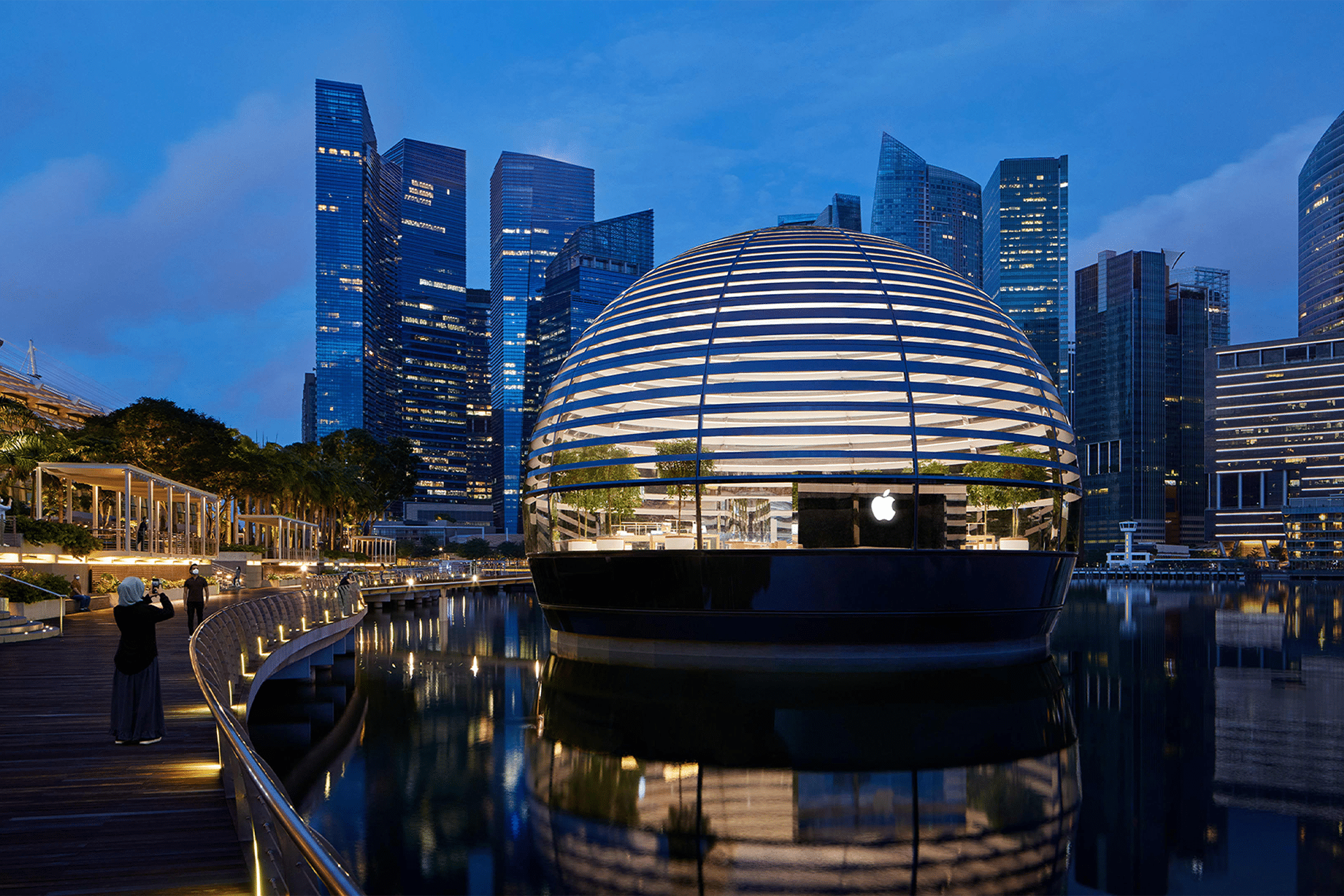 Marina Bay - A round, glass-domed Apple Store near me floats on Marina Bay in Singapore at dusk, illuminated with horizontal white lines and surrounded by water, with city skyscrapers and a boardwalk in the background.