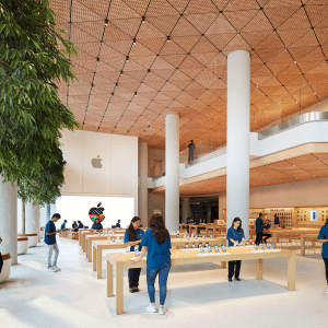 Spacious Apple Store Near Me interior with wooden tables showcasing devices, employees in blue shirts assisting customers, a large tree on the left, and a colorful Apple logo on the back wall. Bright, open modern design with two floors.