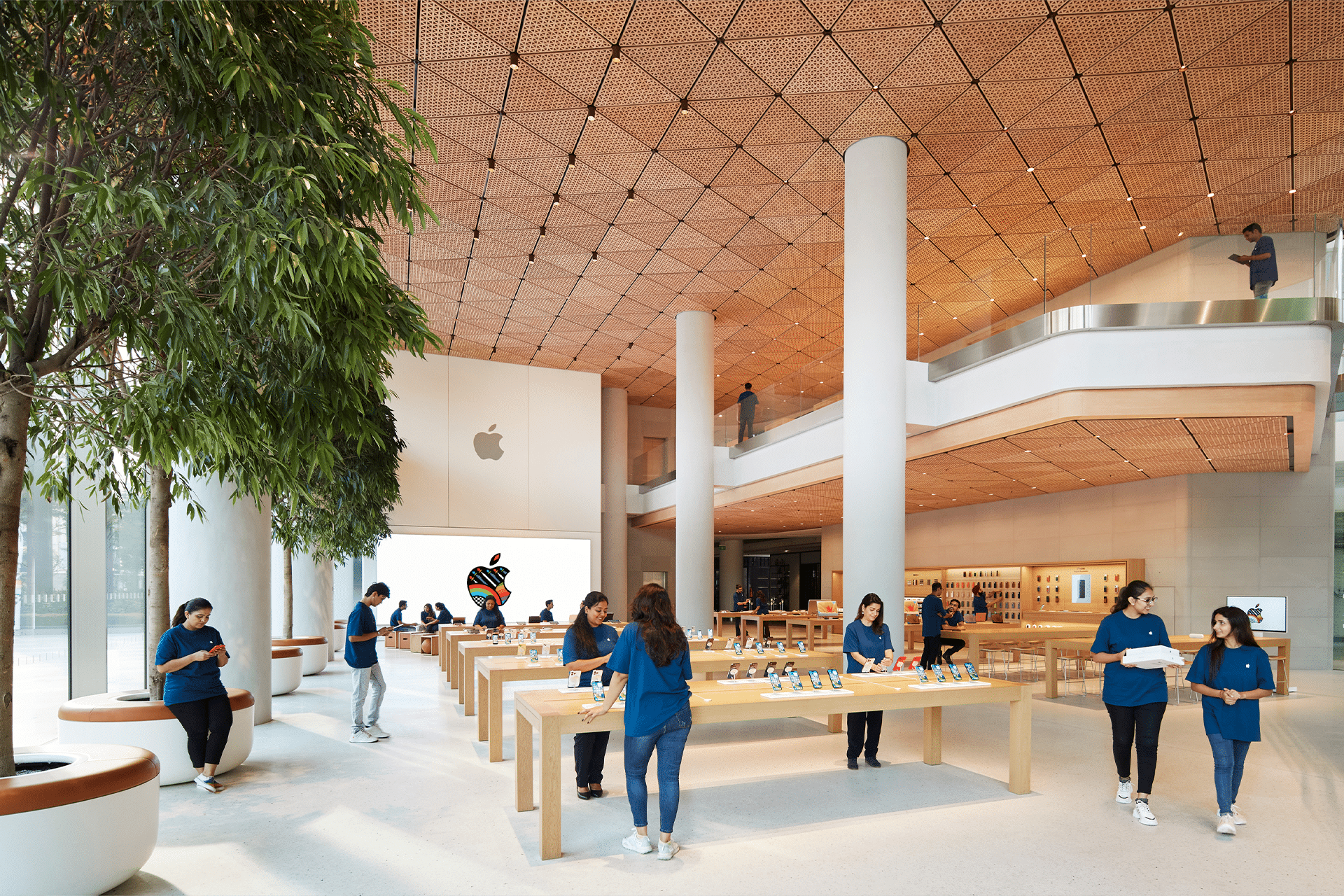 Spacious Apple Store Near Me interior with wooden tables showcasing devices, employees in blue shirts assisting customers, a large tree on the left, and a colorful Apple logo on the back wall. Bright, open modern design with two floors.