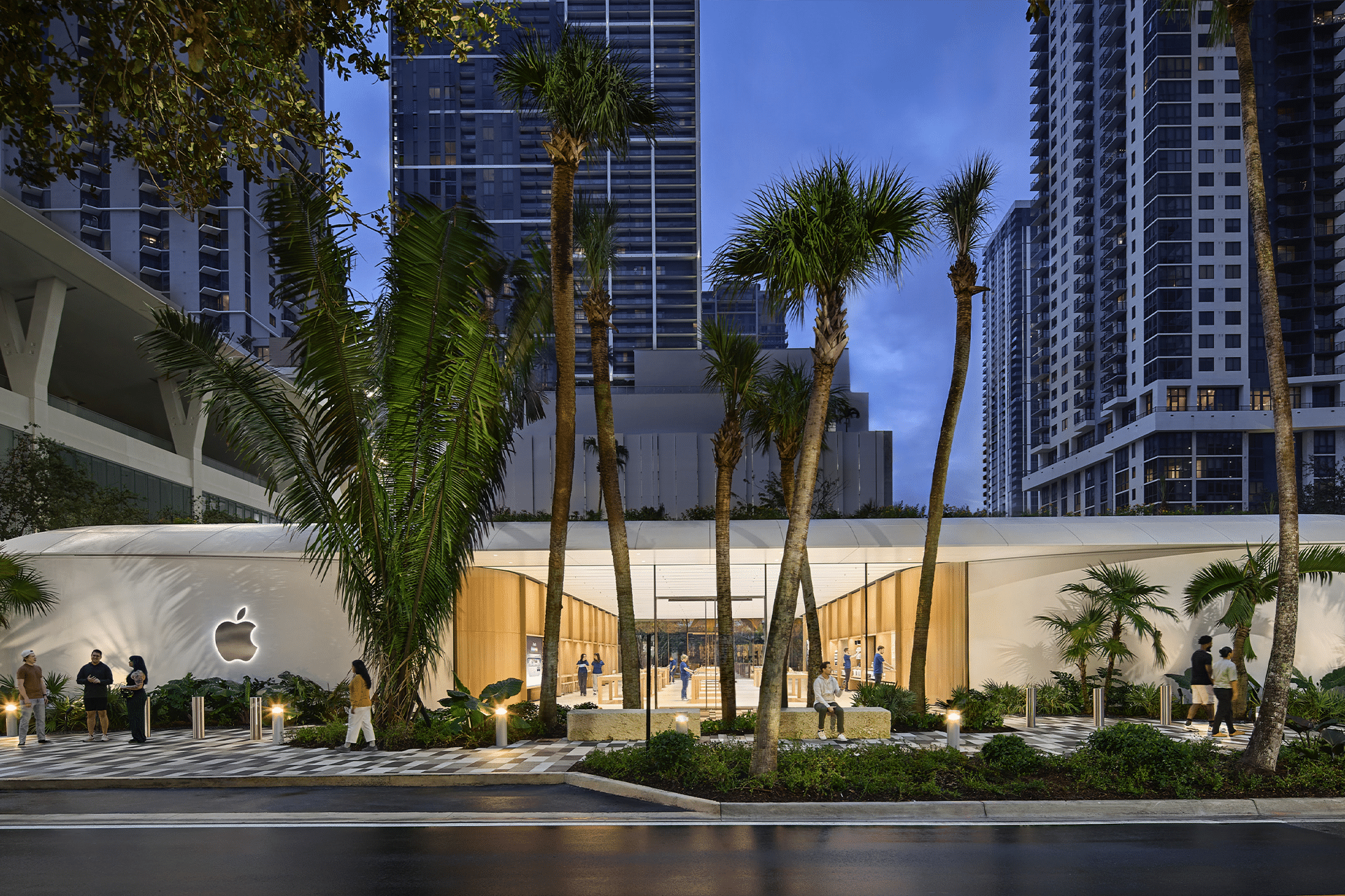 A modern Apple Store near me features a white curved roof and glass walls, nestled among palm trees and tall city buildings at dusk, with people strolling by the entrance.