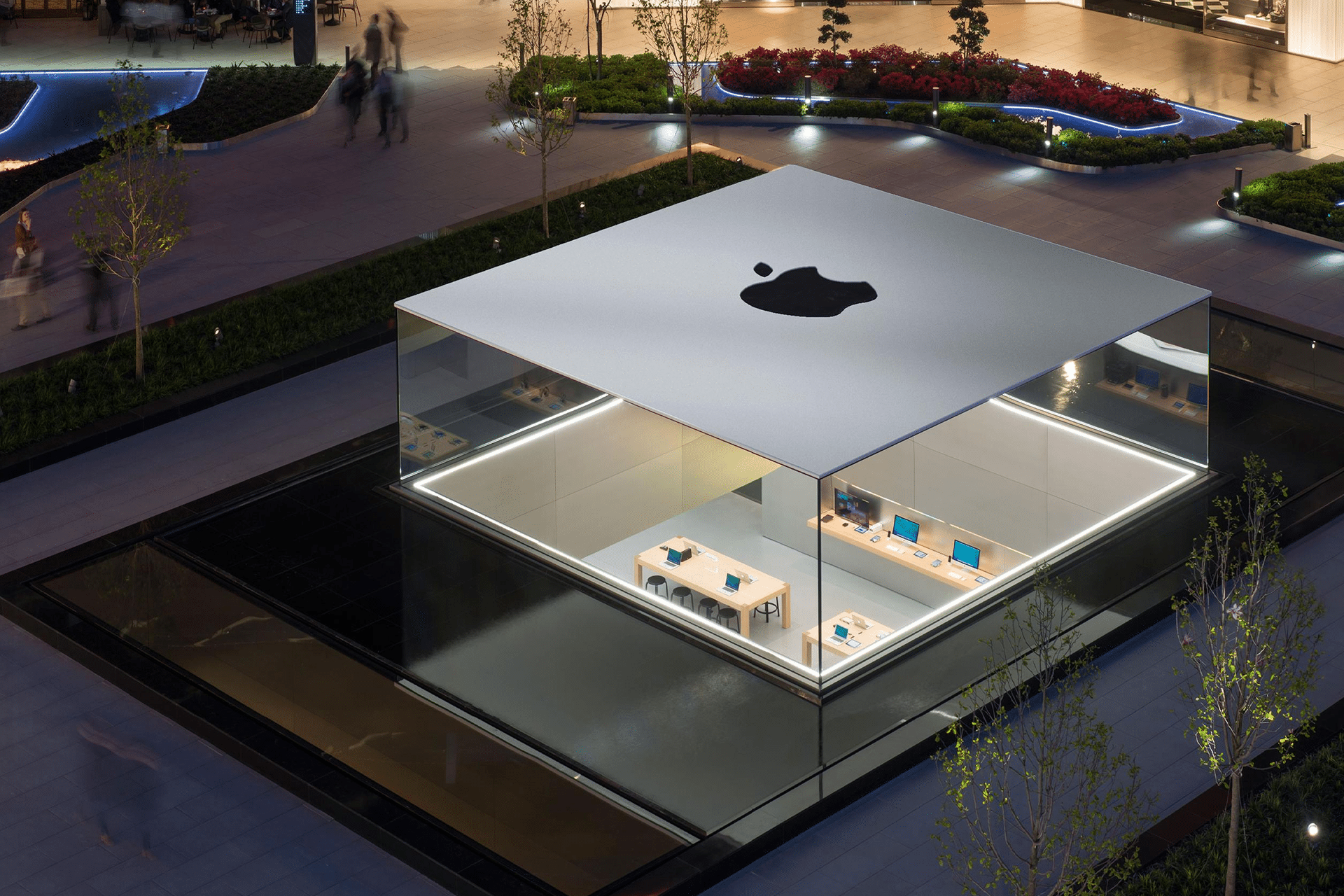 A modern, glass-walled Apple Store near me sits surrounded by water and landscaped greenery, with tables displaying Apple products inside. The building has a large Apple logo on the flat, white roof. People walk nearby.