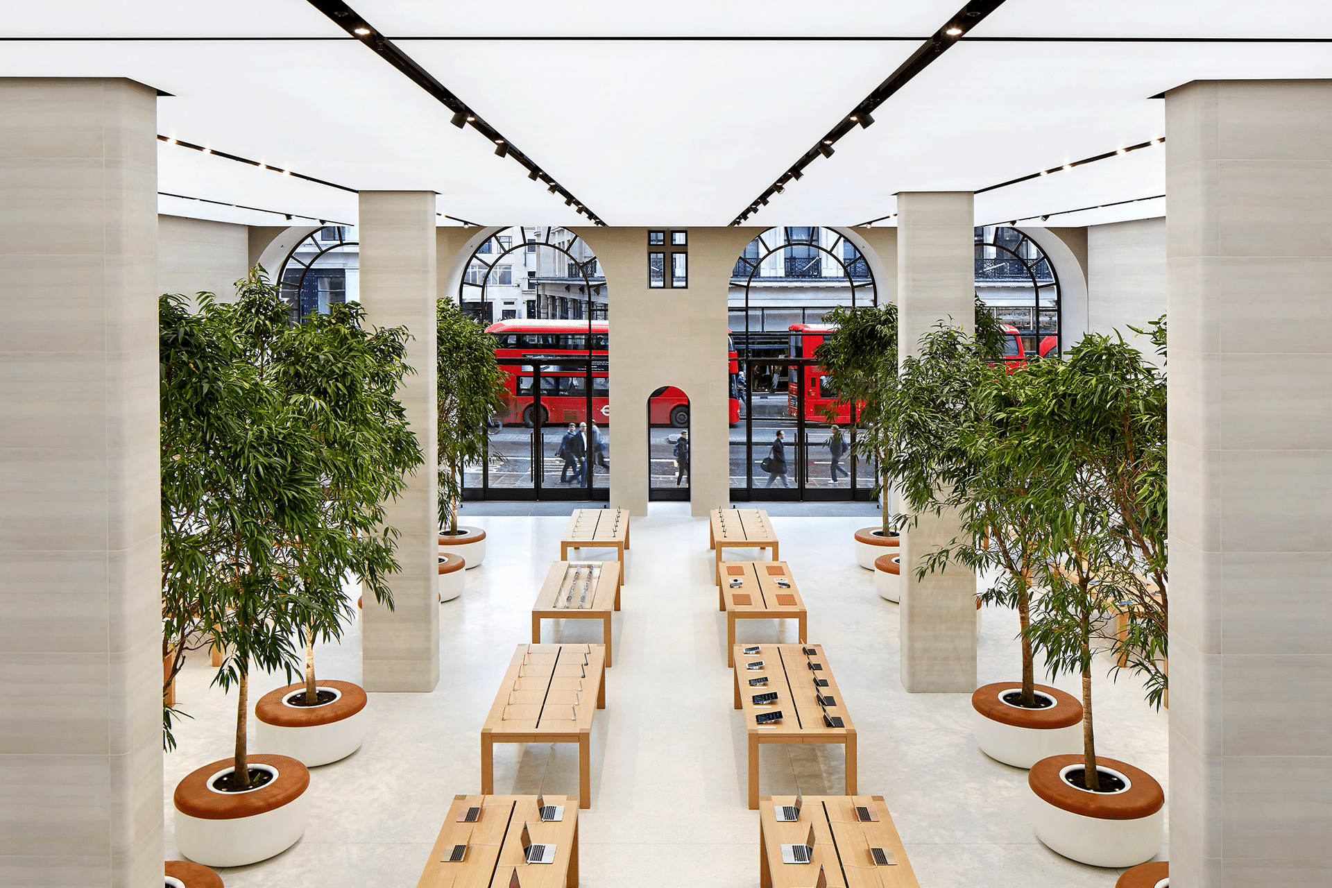 Apple market cap - A spacious, modern Apple Store near me features large potted trees, wooden tables displaying electronics, tall columns, and arched windows. Red double-decker buses are visible outside through the windows.
