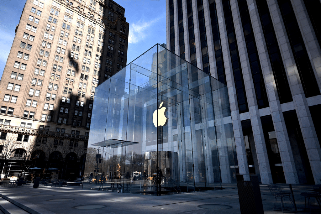 A large glass cube with an illuminated Apple logo marks the entrance to an Apple Store near me, surrounded by tall buildings under a clear blue sky.
