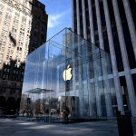 A large glass cube with an illuminated Apple logo marks the entrance to an Apple Store near me, surrounded by tall buildings under a clear blue sky.