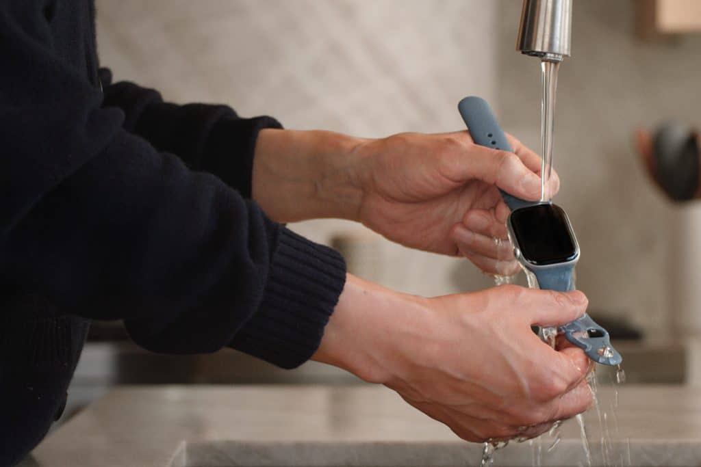 Apple Watch Cleaning - A person demonstrates Apple Watch cleaning by rinsing the smart watch under running water from a kitchen faucet, holding it over a sink with both hands.
