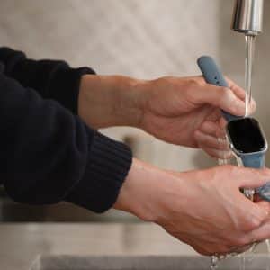 Apple Watch Cleaning - A person demonstrates Apple Watch cleaning by rinsing the smart watch under running water from a kitchen faucet, holding it over a sink with both hands.