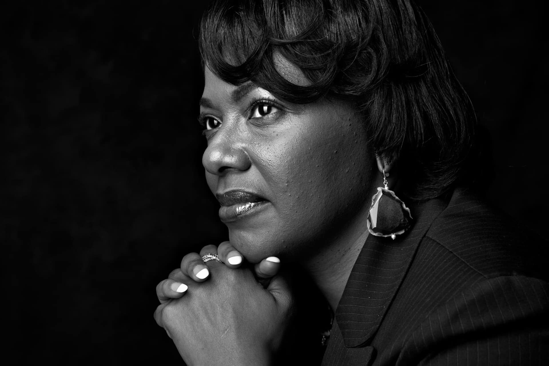 Black and white portrait of a woman in profile, resting her chin on her clasped hands. She wears a suit jacket and large earrings, gazing thoughtfully into the distance, evoking the contemplative spirit of Martin Luther King against a dark background.