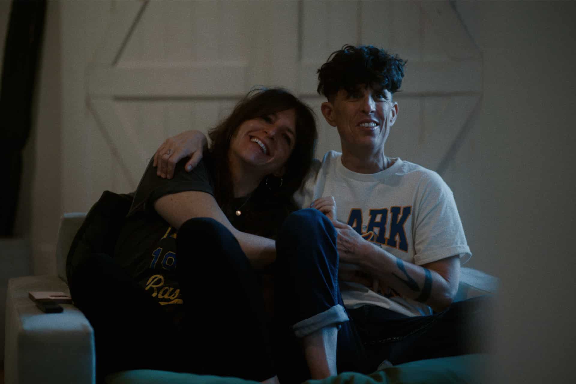 Two people sit closely together on a couch, smiling and laughing as they explore Apple Creator Studio. One with long hair leans on the other, who has short hair. Both appear relaxed and happy in a cozy indoor setting.