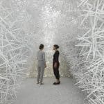 Two people stand facing each other in a room filled with dense, white, crisscrossing sticks forming walls and ceiling, creating an abstract, maze-like environment reminiscent of computational art.