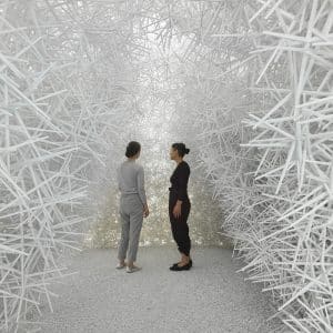 Two people stand facing each other in a room filled with dense, white, crisscrossing sticks forming walls and ceiling, creating an abstract, maze-like environment reminiscent of computational art.