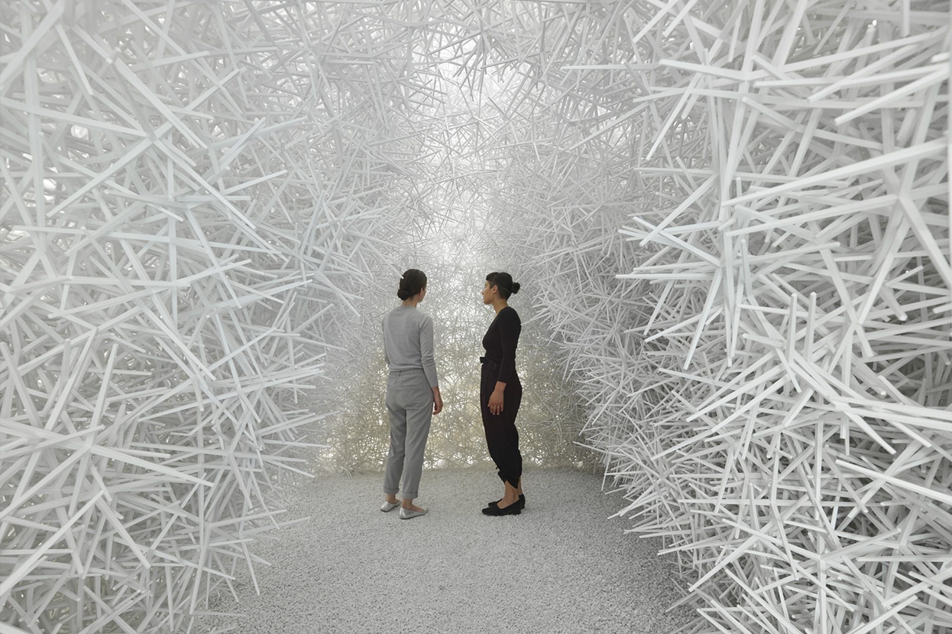 Two people stand facing each other in a room filled with dense, white, crisscrossing sticks forming walls and ceiling, creating an abstract, maze-like environment reminiscent of computational art.