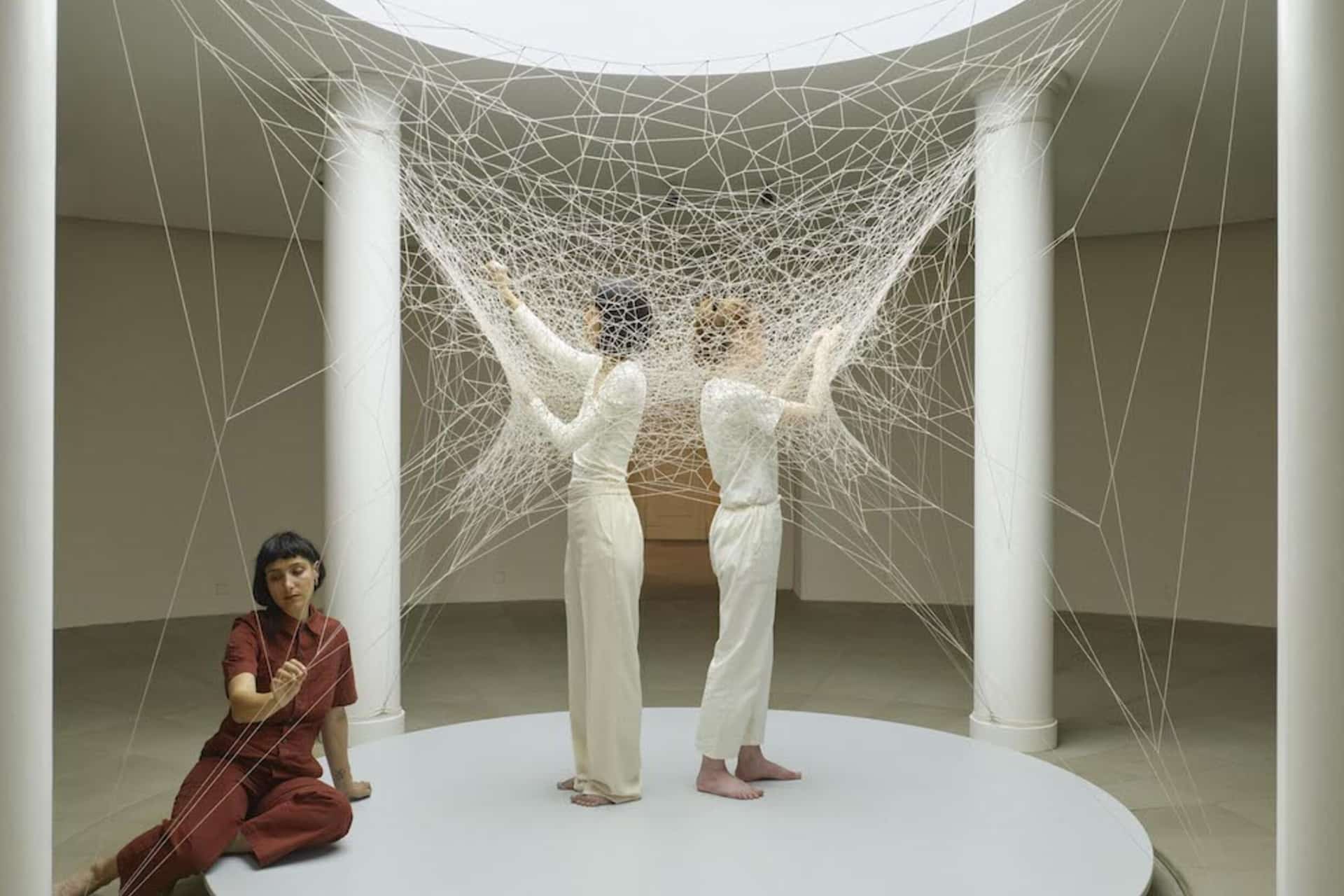Three women engage with a large, intricate web of white string—reminiscent of Computational Art—suspended between white columns in a round, minimalist space. Two stand back to back inside the web while one sits outside, holding a strand.