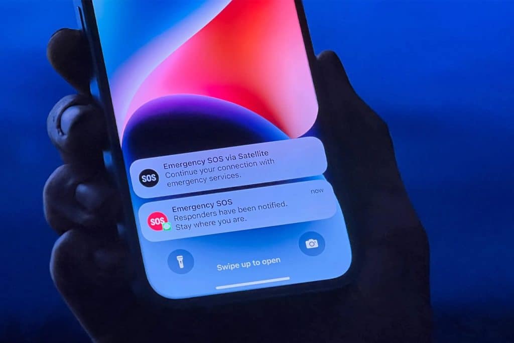 iPhone held in a hand displaying Emergency SOS via Satellite and iPhone Crash Detection alerts on the lock screen.