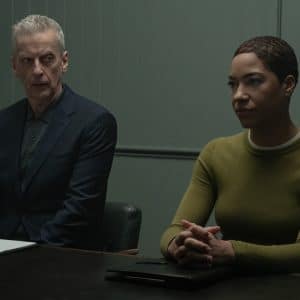 Two people sit at a table in a dimly lit room, reminiscent of an Apple TV Criminal Record scene. The man wears a dark suit with a folder before him, while the woman in a green sweater sits with her hands clasped on the table.