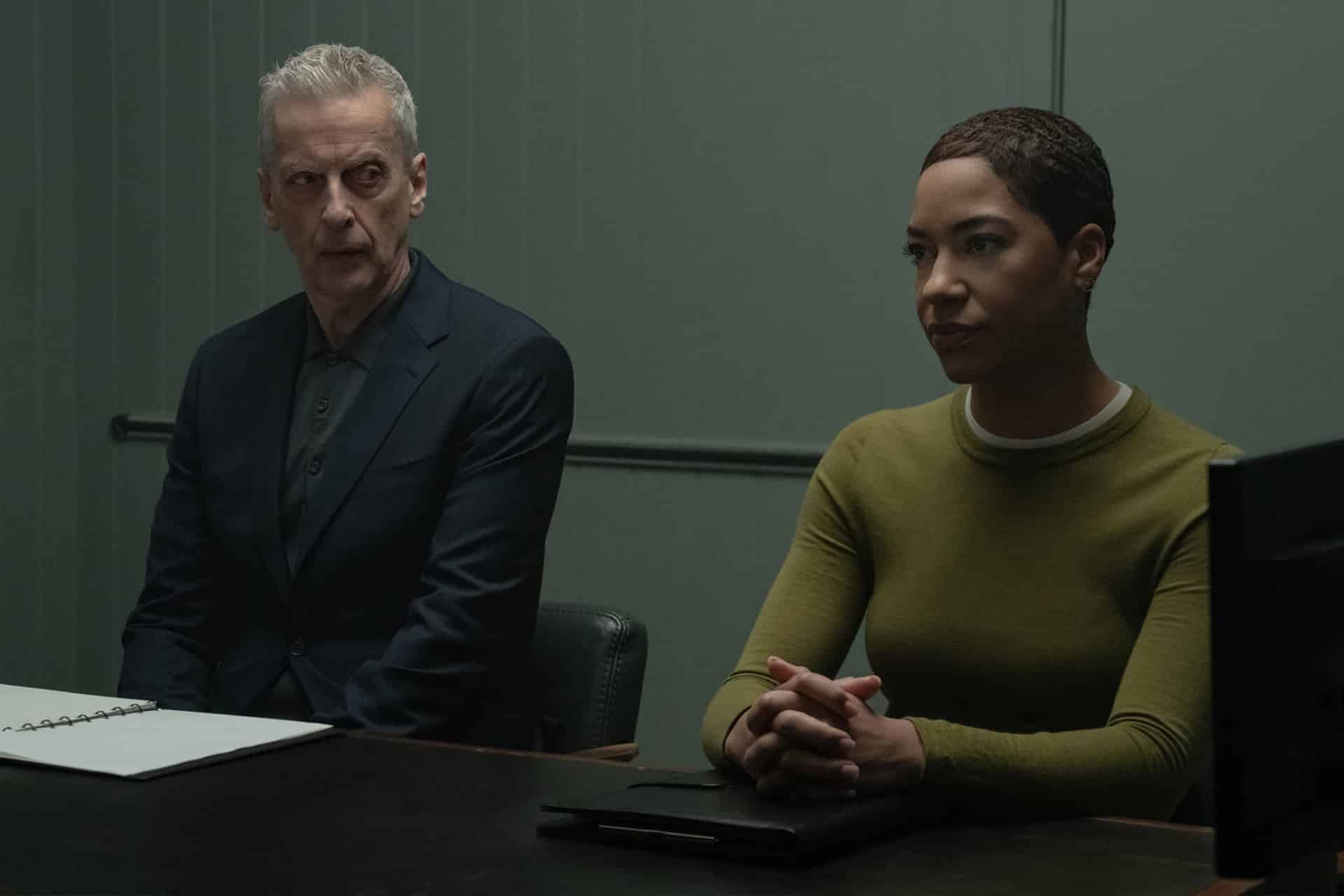 Two people sit at a table in a dimly lit room, reminiscent of an Apple TV Criminal Record scene. The man wears a dark suit with a folder before him, while the woman in a green sweater sits with her hands clasped on the table.