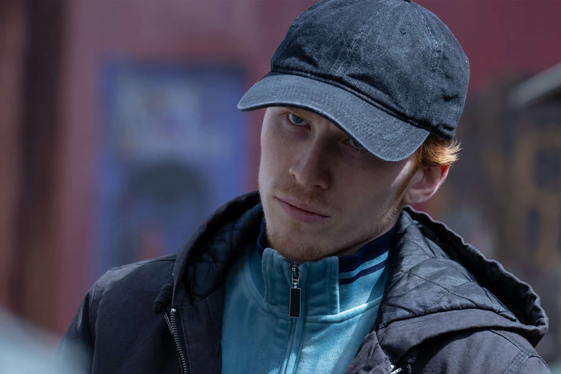 A young man with light skin, red hair, and a short beard wears a black cap and blue jacket. He looks seriously at the camera, standing outdoors with a blurred urban background—like a scene from an Apple TV Criminal Record episode.