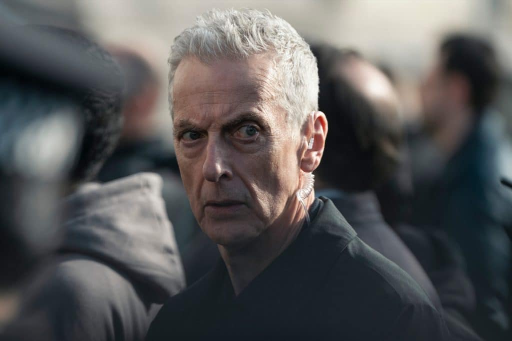 A man with short gray hair and a serious expression stands in a crowd, looking directly at the camera. He is wearing a dark jacket and an earpiece—reminiscent of intense scenes from Apple TV shows like Criminal Record Season 2.
