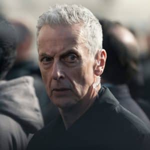 A man with short gray hair and a serious expression stands in a crowd, looking directly at the camera. He is wearing a dark jacket and an earpiece—reminiscent of intense scenes from Apple TV shows like Criminal Record Season 2.