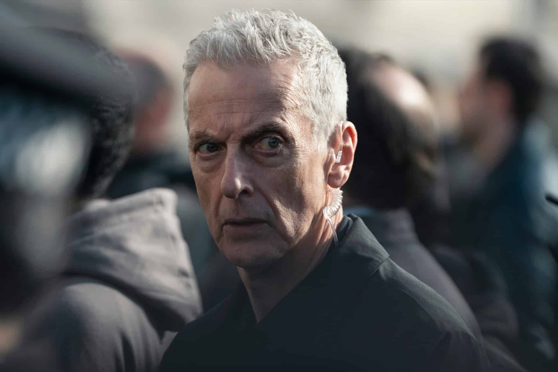 A man with short gray hair and a serious expression stands in a crowd, looking directly at the camera. He is wearing a dark jacket and an earpiece—reminiscent of intense scenes from Apple TV shows like Criminal Record Season 2.