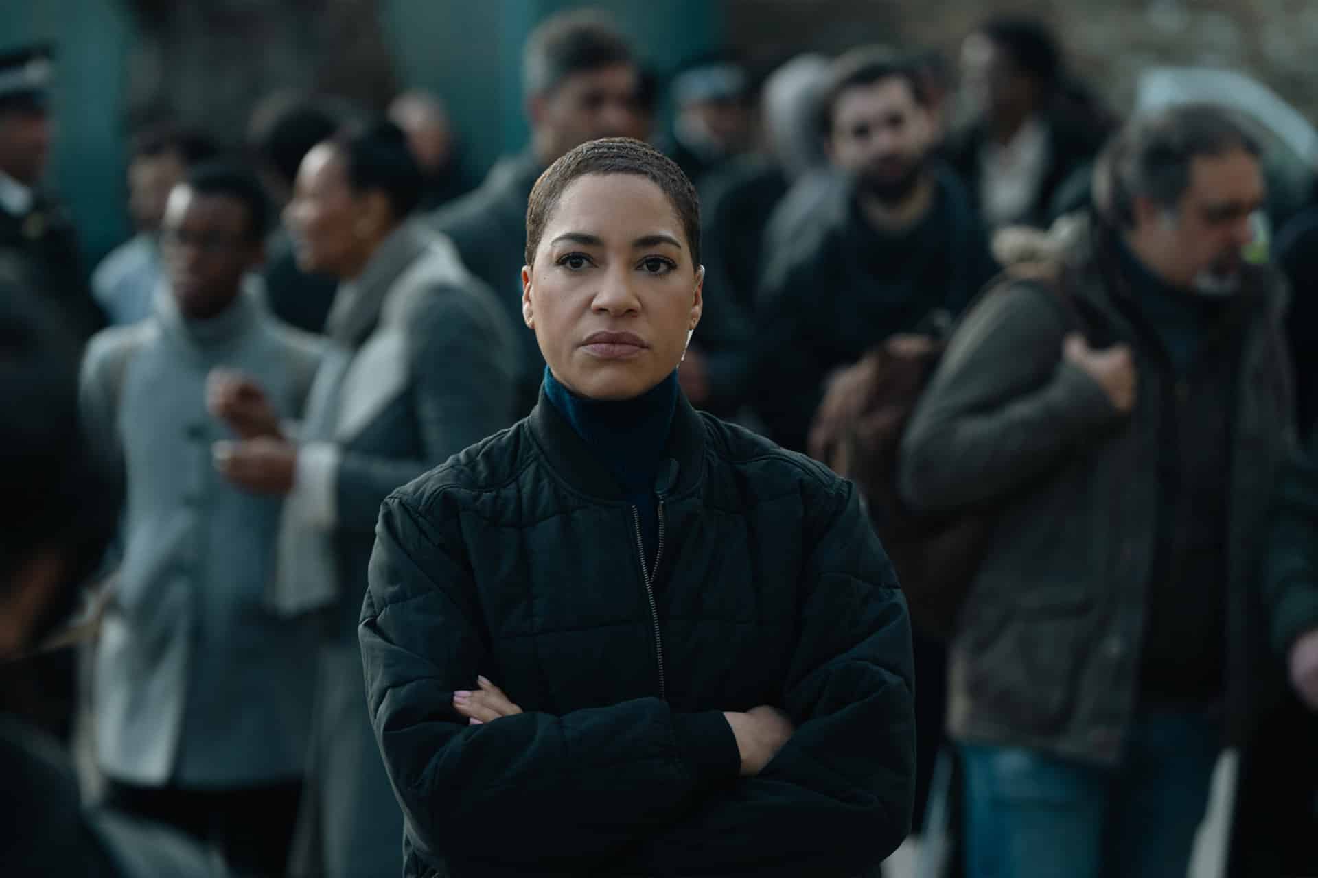 A woman with short hair and a serious expression stands with her arms crossed in the foreground, reminiscent of intense moments seen in Apple TV Criminal Record, while a blurry crowd gathers behind her outdoors.