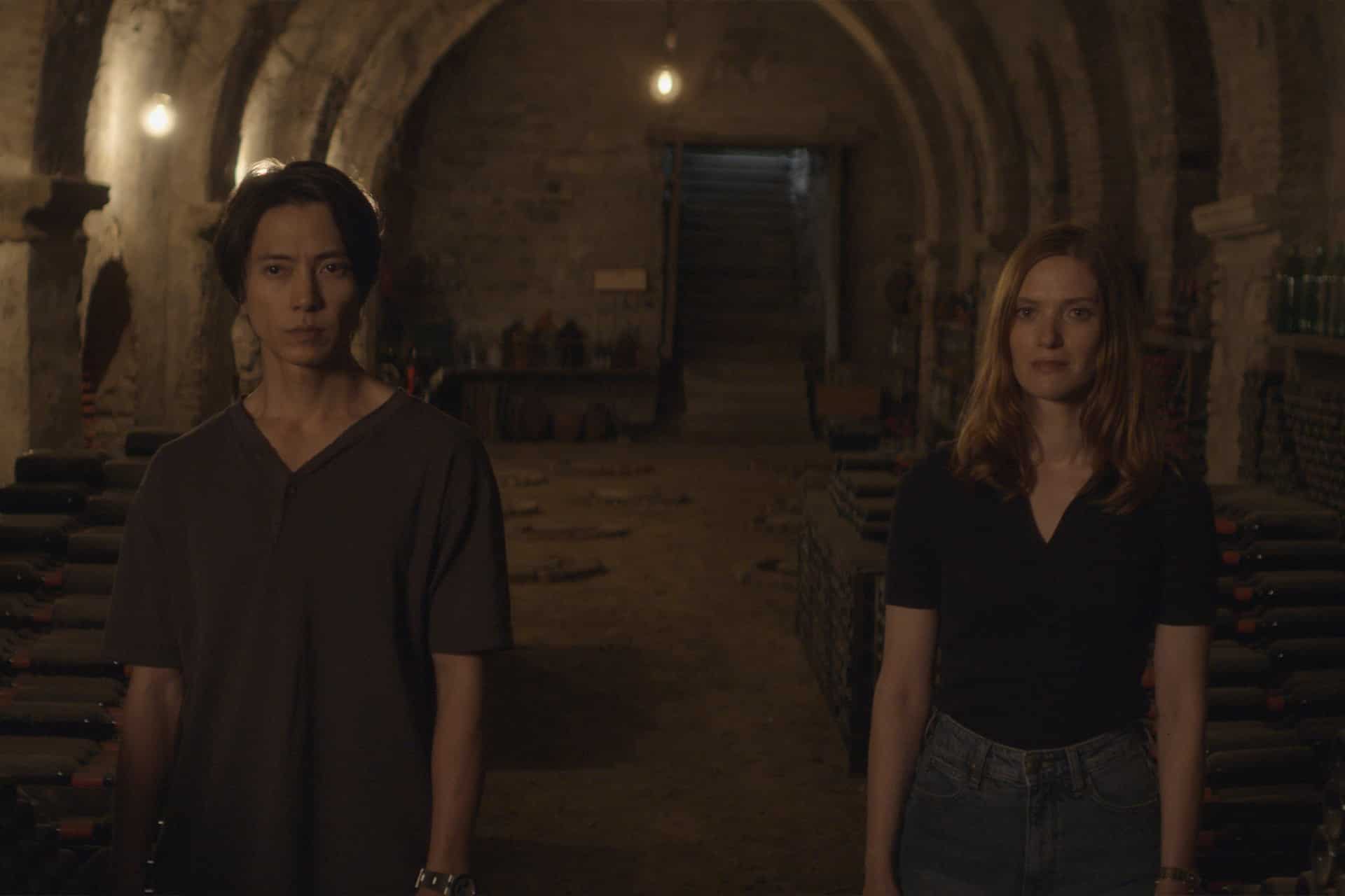 A man and a woman stand side by side in a dimly lit cellar, surrounded by shelves of wine bottles, evoking the intrigue of Apple TV Drops of God, with stone arches and stairs leading up at the back.