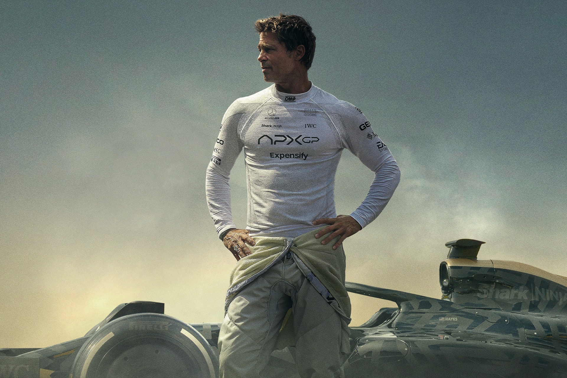 A man in a white racing suit stands confidently before a Formula 1 car, his suit unzipped to the waist and hands on hips, gazing to the side as dramatic clouds loom—captured with cinematic flair in Apple Creator Studio.