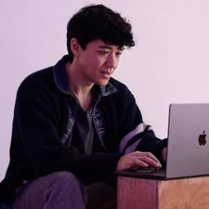 A person with dark, curly hair sits on a wooden block, focused on their laptop with an Apple logo as they edit in Final Cut Pro. They wear a dark cardigan with a striped sleeve over a dark shirt, set against a pale purple background.