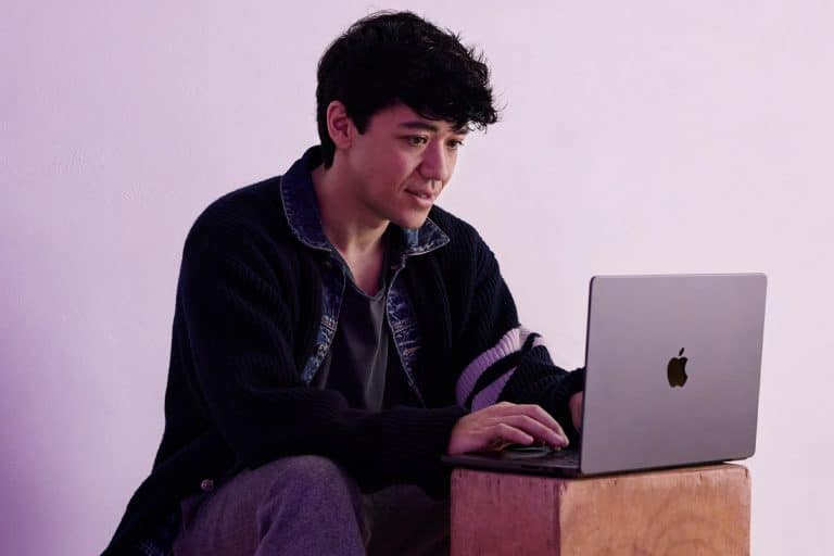 A person with dark, curly hair sits on a wooden block, focused on their laptop with an Apple logo as they edit in Final Cut Pro. They wear a dark cardigan with a striped sleeve over a dark shirt, set against a pale purple background.