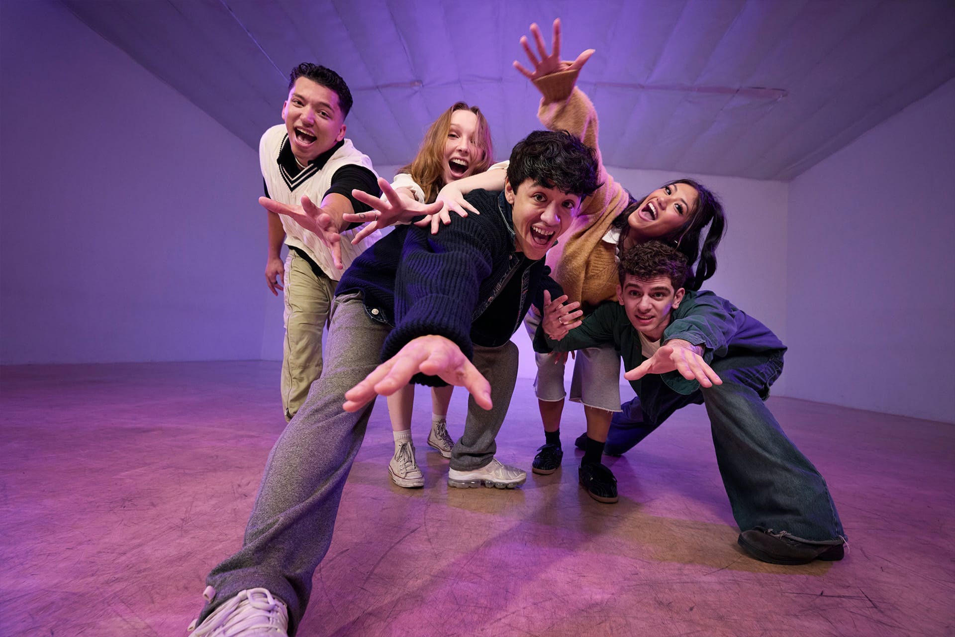 Five young adults enthusiastically pose together indoors, reaching toward the camera with big smiles and dynamic energy under purple lighting—perfect for capturing a vibrant group moment to edit later in Final Cut Pro.