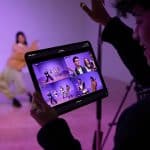 A person holds a tablet displaying multiple video feeds of two people dancing in a studio, while filming the dancers with a camera on a tripod. The purple-lit background hints at post-production editing in Final Cut Pro.
