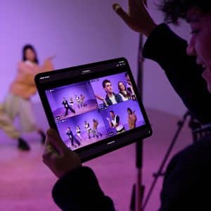 A person holds a tablet displaying multiple video feeds of two people dancing in a studio, while filming the dancers with a camera on a tripod. The purple-lit background hints at post-production editing in Final Cut Pro.