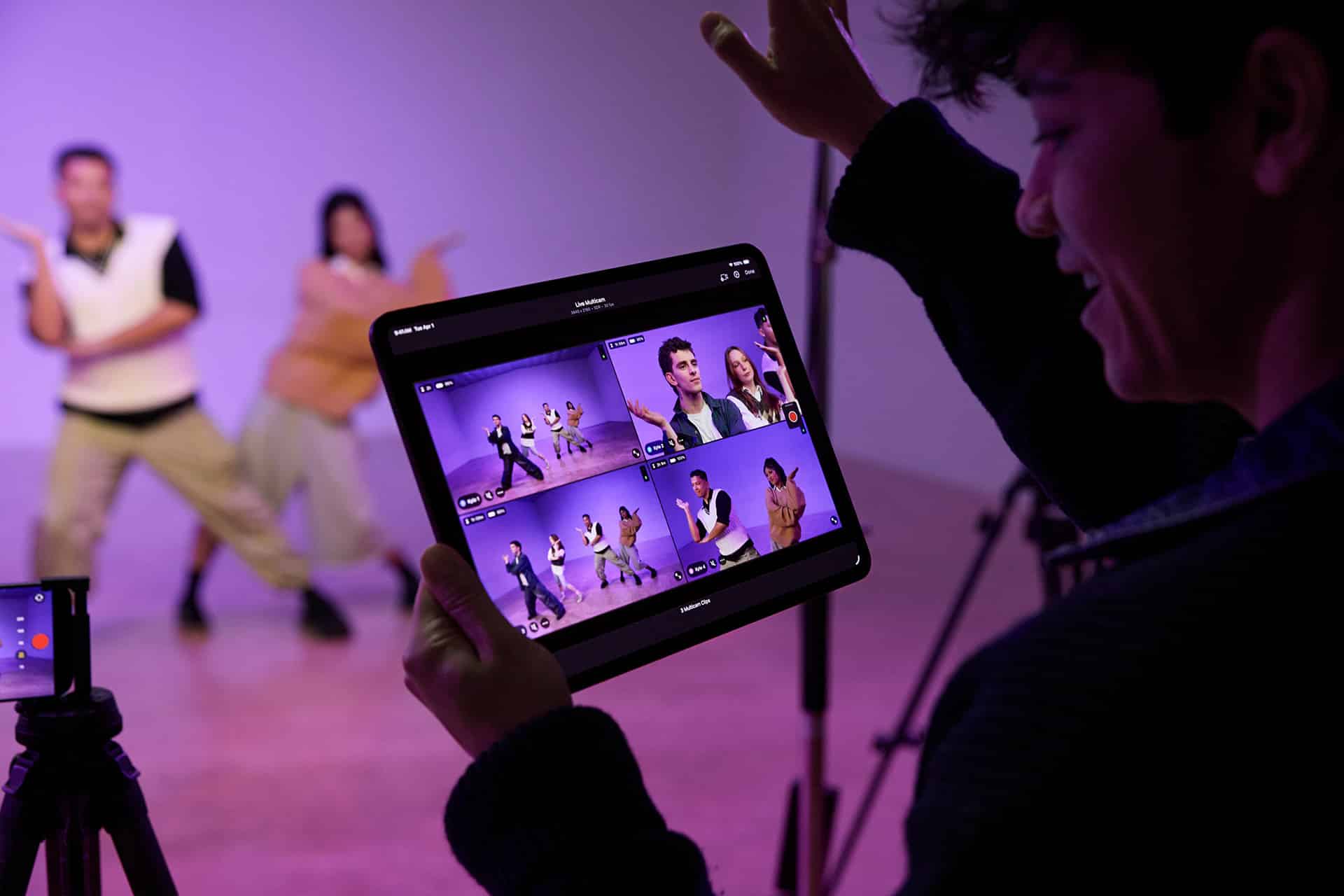 A person holds a tablet displaying multiple video feeds of two people dancing in a studio, while filming the dancers with a camera on a tripod. The purple-lit background hints at post-production editing in Final Cut Pro.