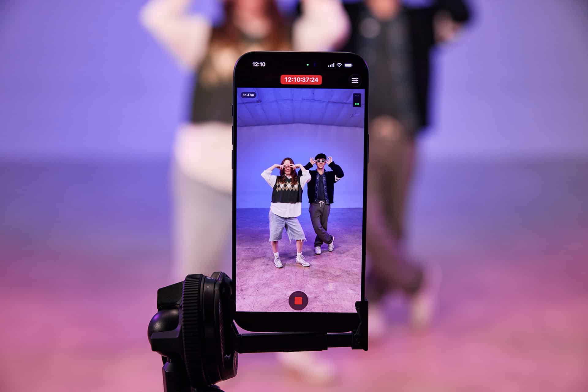 A smartphone on a tripod records two people dancing indoors. The phone screen, ready for Final Cut Pro editing, shows both dancers posing in a well-lit, purple-toned room, while the figures appear blurred in the background.