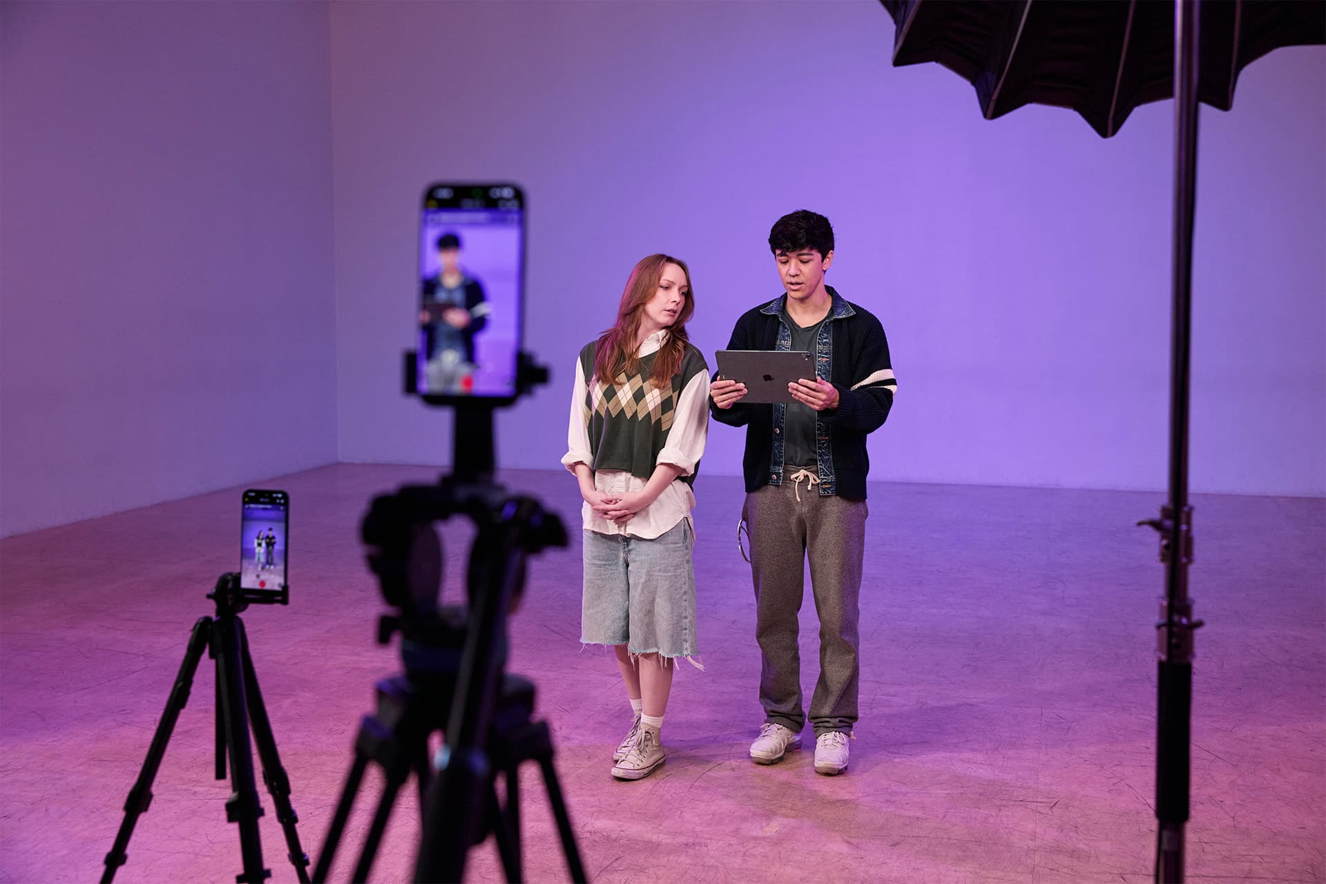 Two people stand in a studio with purple lighting, reading from a tablet. A smartphone on a tripod records them for editing later in Final Cut Pro, while a large studio light is visible overhead. The background is plain and the floor is bare.