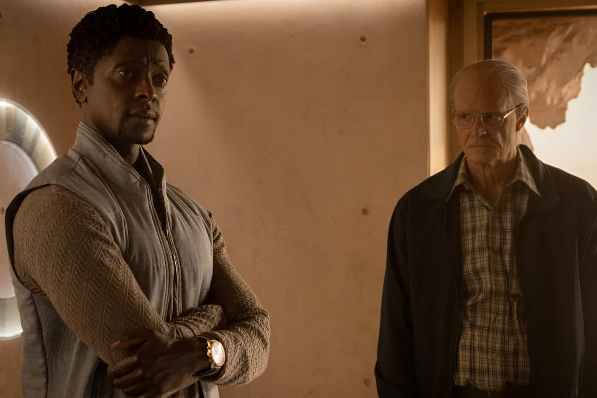 Two men stand indoors; the man on the left has dark skin and curly hair, wearing a light gray vest with arms crossed, while the older man on the right, seen in For All Mankind Season 5, wears glasses and a jacket, looking serious.